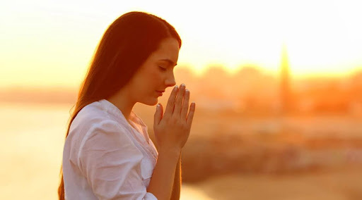 Young woman praying with eyes closed at sunrise, using spiritual connection to overcome stress.