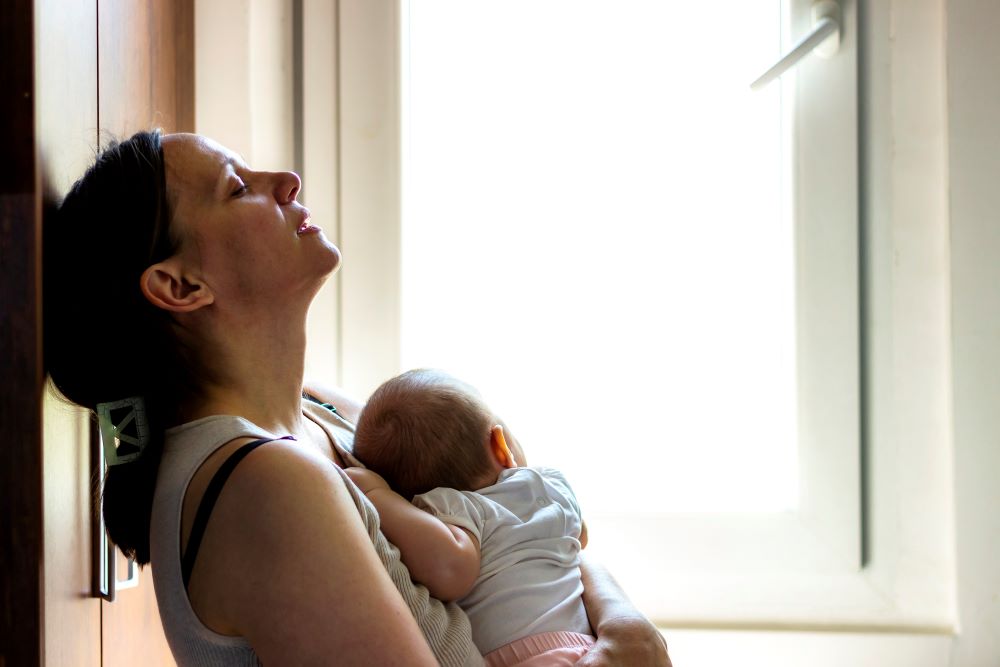 Exhausted mother holding baby experiencing postpartum depression near a window, seeking Christian counseling support for healing and peace.